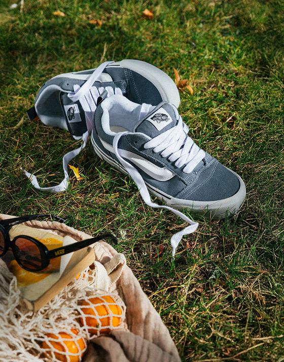 A pair of gray and white Knu Skool lie in the grass with their laces untied next to a grocery bag for a picnic.