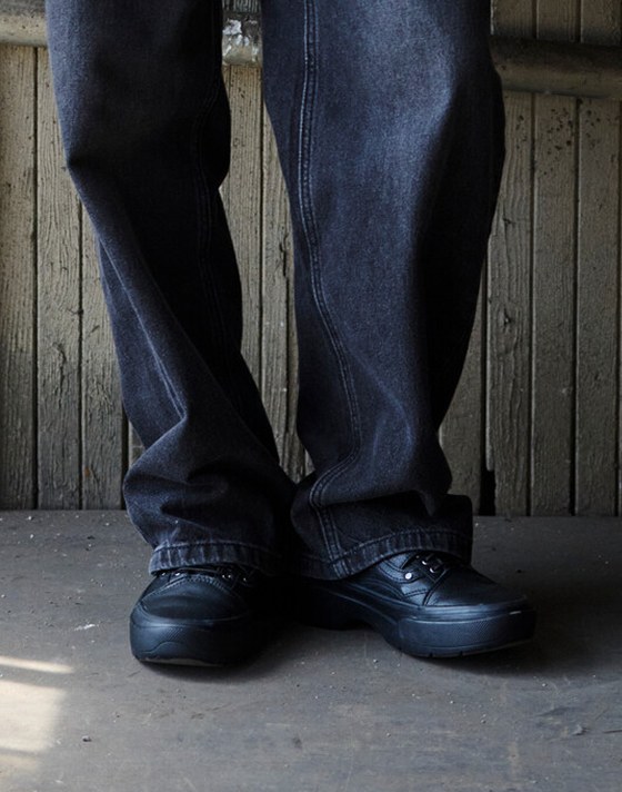 A knee-down shot of someone wearing black-on-black Premium Old Skool with black jeans in a warehouse.