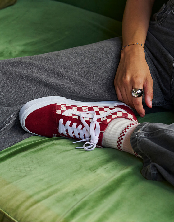 A close-up of a red Checkerboard Old Skool paired with gray jeans on a green felt couch.
