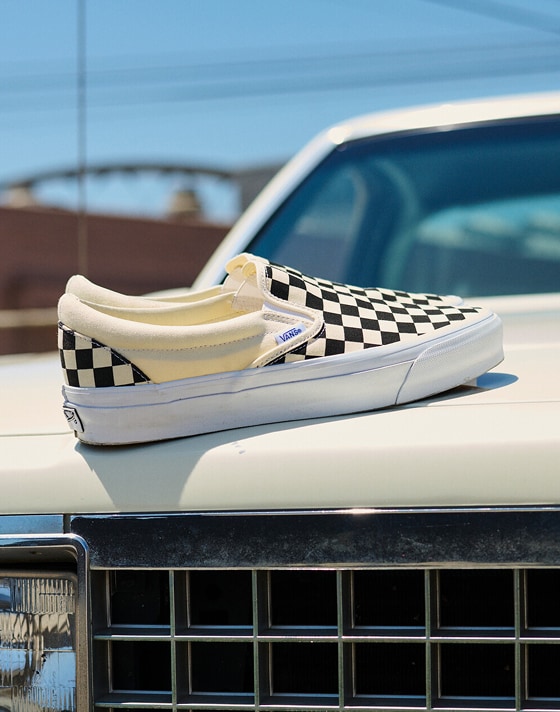 A pair of black and white Checkerboard Classic Slip-On rest on the hood of an old white car under the sun.