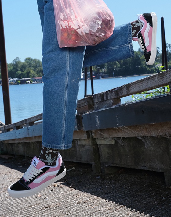 A young woman jumps on a boat ramp near the water wearing pink and black Knu Skool with blue jeans.