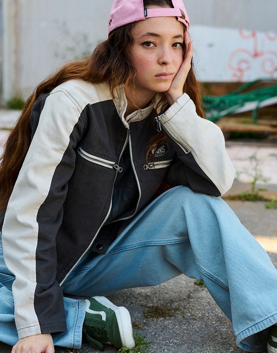 A young woman sits on the asphalt wearing a Ryden Biker Jacket, Cinched Check 5 Baggy blue jeans, and pink Check Side Curved Bill Jockey Hat.