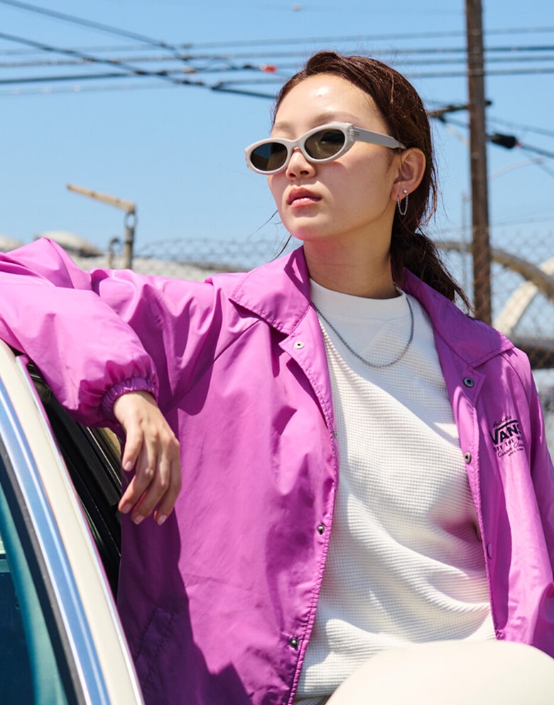 A woman leans against a Chevy Camino wearing a white Navigate Long Sleeve Thermal, a purple Vans jacket, and sunglasses.