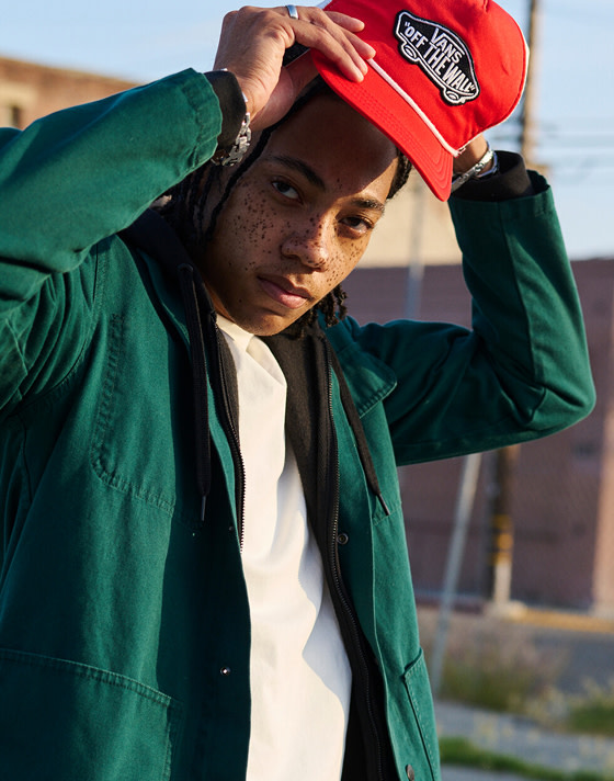 A man looks into the camera fixing his red Vans “Off the Wall” hat paired with a green overshirt.