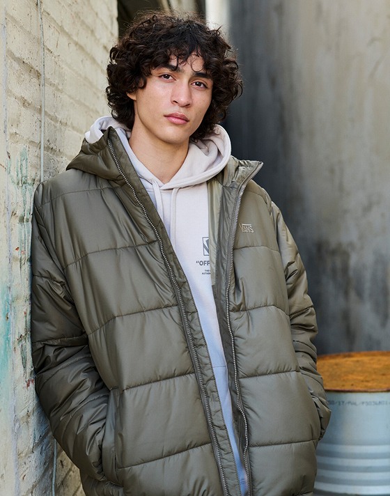 A man poses in a warehouse and looks into the camera wearing a taupe mist Stockpile Pullover Hoodie and brown MTE Hillgate Puffer.