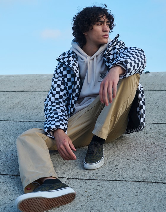 A young man sits on the roof of a warehouse wearing a Stockpile Pullover, Range Relaxed Elastic Pants, Skate Authentic, and a Checkerboard jacket.