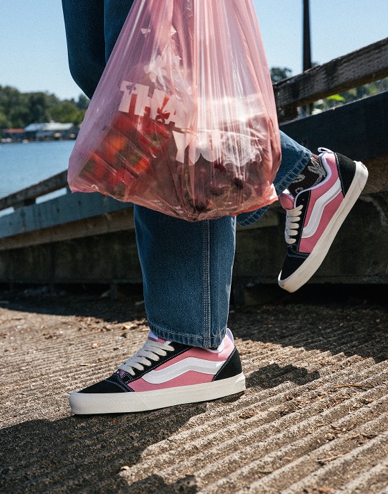 A knee-down shot of someone wearing pink and black Knu Skool with blue jeans on a boat ramp near the water.
