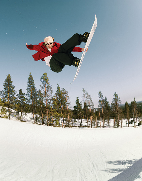A snowboarder jumps off a roof wearing Vans clothes and snowboarding boots in a grainy black and white photo.