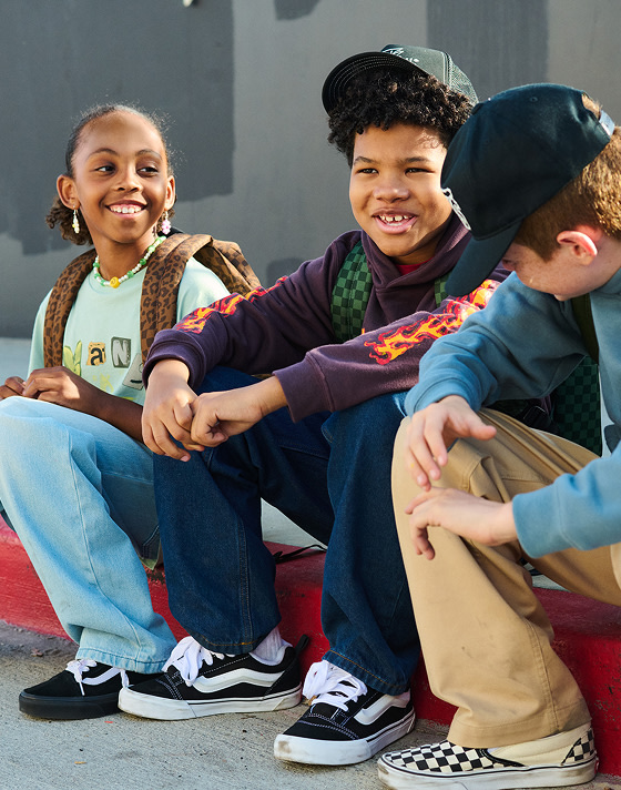 Three young kids sit on a curb wearing Vans clothes, backpacks, Old Skool, Knu Skool, and Checkerboard Classic Slip-On.