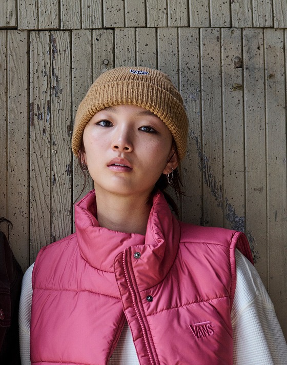 A woman poses outside a warehouse with a mauvewood MTE Hillgate Cropped Vest and Hex Patch Cuff Beanie.