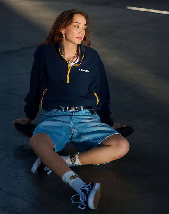 A young woman sits on a concrete floor wearing denim shorts, white Vans crew socks, and black and white Old Skool.