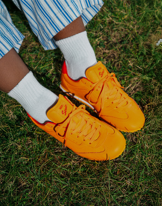 A close-up of someone wearing solar orange Super Lowpro with paired white socks and blue-striped pants on the grass.