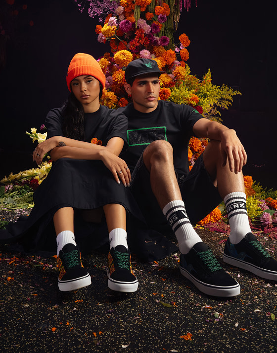 Two people sit in front of a display of marigolds and ferns wearing Vans Día de Muertos tees, beanies, and shoes.