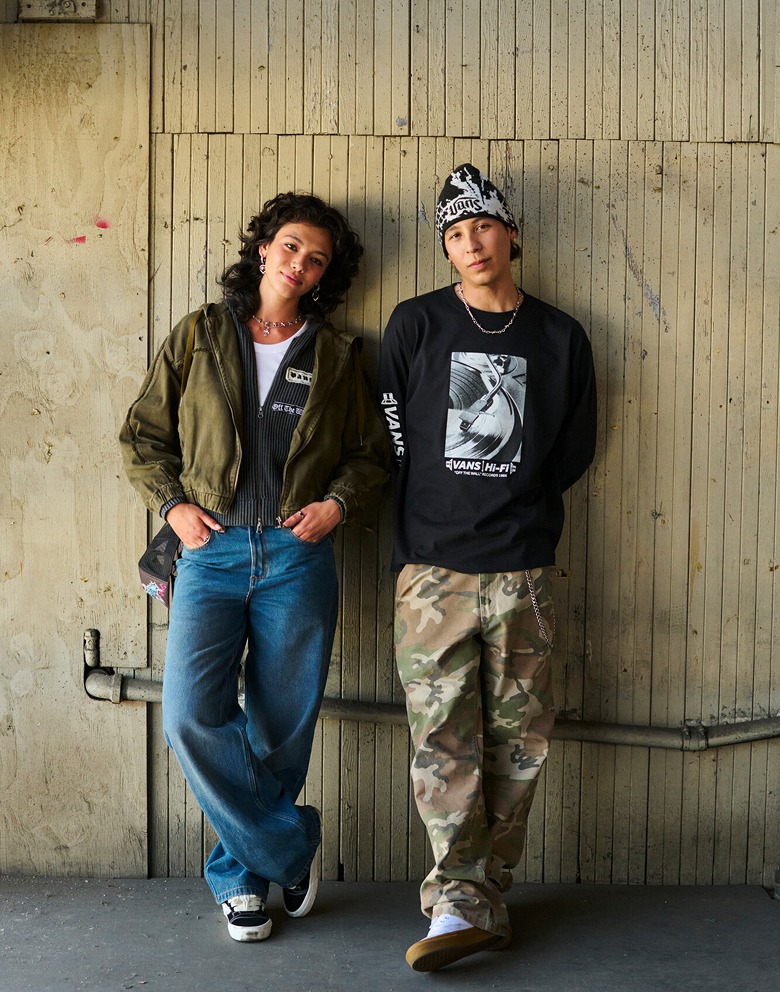 A young woman and man lean up against a wood-paneled wall outside of a warehouse wearing Vans clothes.