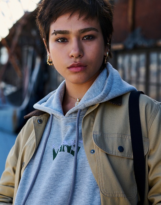 A young woman with short hair and a septum piercing looks at the camera wearing a gray Vans hoodie and a tan Vans jacket.