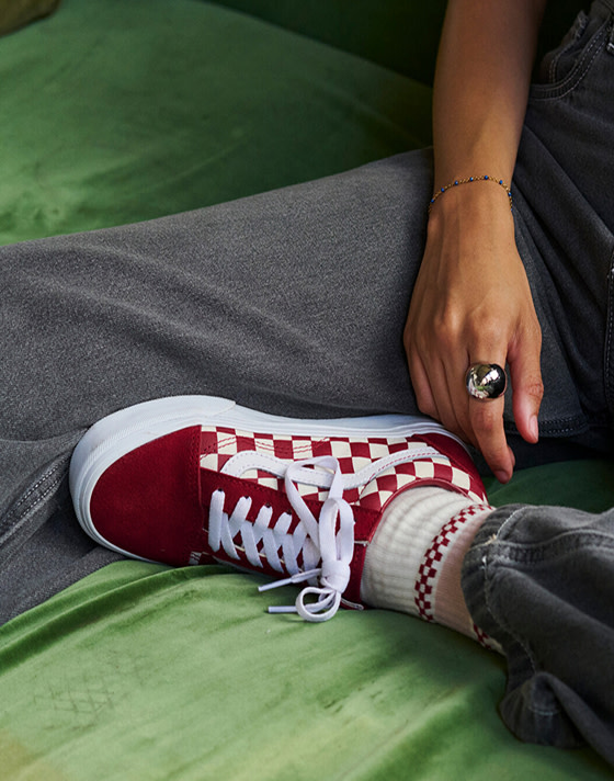 A close-up of a red Checkerboard Old Skool paired with gray jeans on a green felt couch.