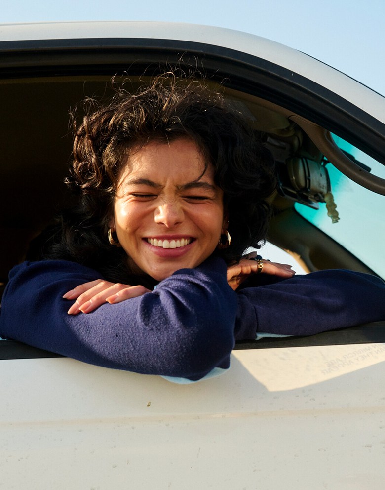 A young woman sticks her head out of a minivan window and smiles at the camera with her eyes closed on a sunny day.