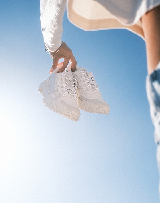 Someone holds a pair of white Crosspath XC GORE-TEX against a blue sky with the sun peeking in.