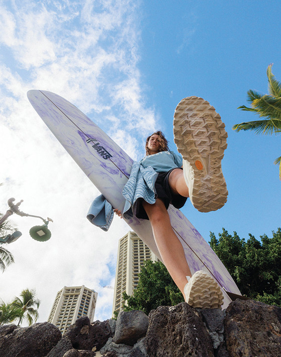 A woman holding a surfboard stands on a rock near a beach and sticks her white Crosspath XC GORE-TEX towards the camera.