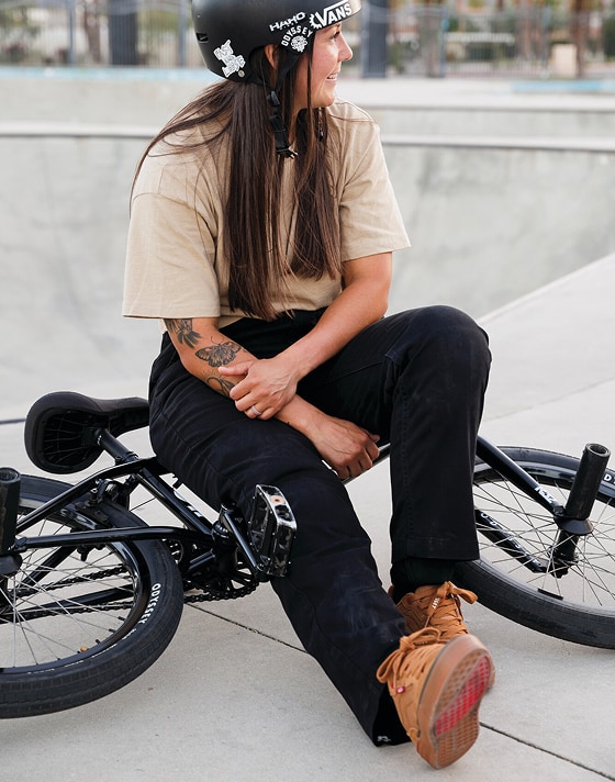 Vans BMX Rider Perris Benegas sits on her bike at a skate park wearing black jeans and the brown BMX Proof WaffleCup.