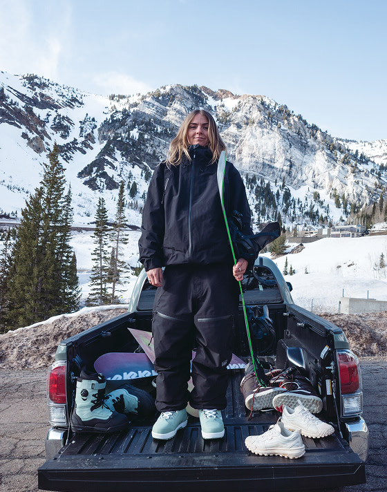 A young woman stands in the bed of a truck wearing all black snowboard gear and the Luna Pro with a mountain in the background.