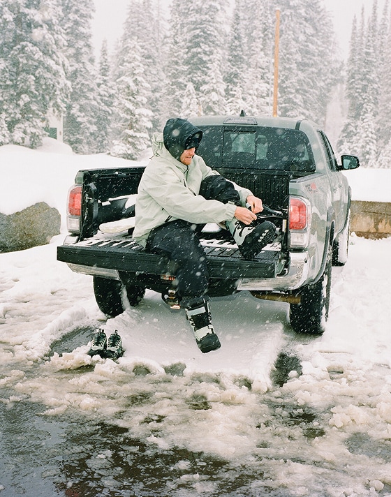 A man puts on the Infuse snowboard boot in the bed of a truck in a snowy parking lot as it snows with frosted pines in the background.
