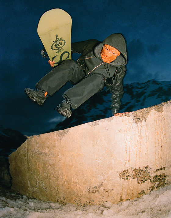 A snowboarder wearing all black and an orange ski mask jumps a wall wearing black Crosspath XC GORE-TEX holding a snowboard.