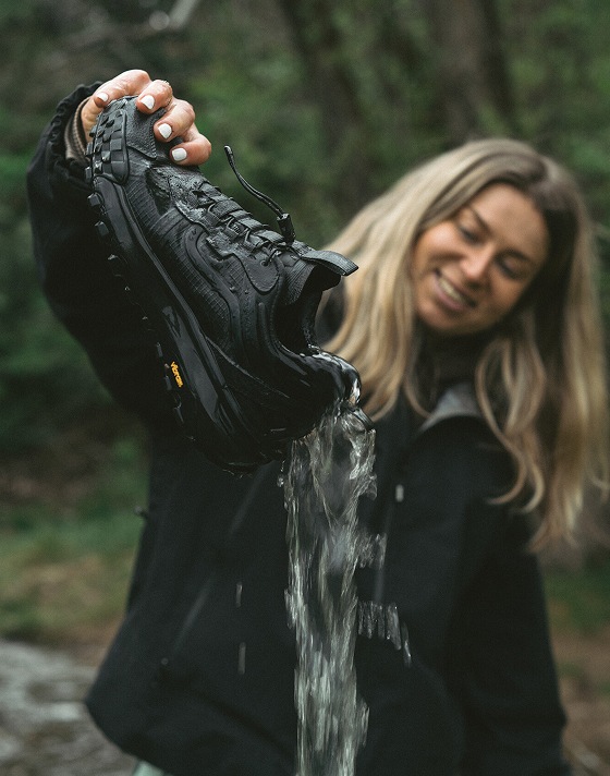 A woman holds a black Crosspath XC GORE-TEX shoe and dumps water out of it next to a river in a forest.