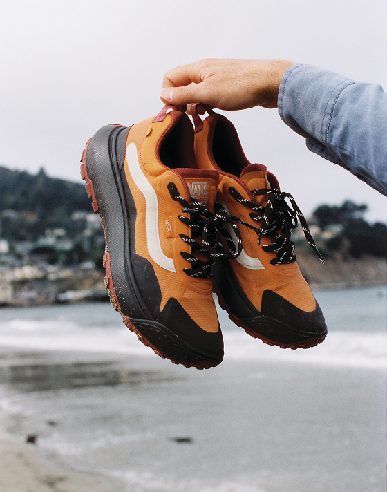 Someone holds up orange Crosspaths by their heel tabs with a foggy beach in the background.