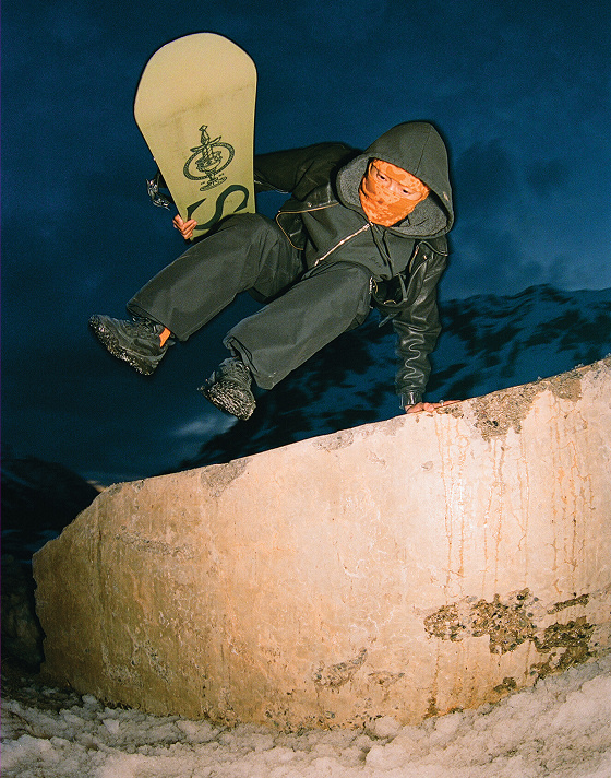 A snowboarder wearing all black and an orange ski mask jumps a wall wearing black Crosspath XC GORE-TEX holding a snowboard.
