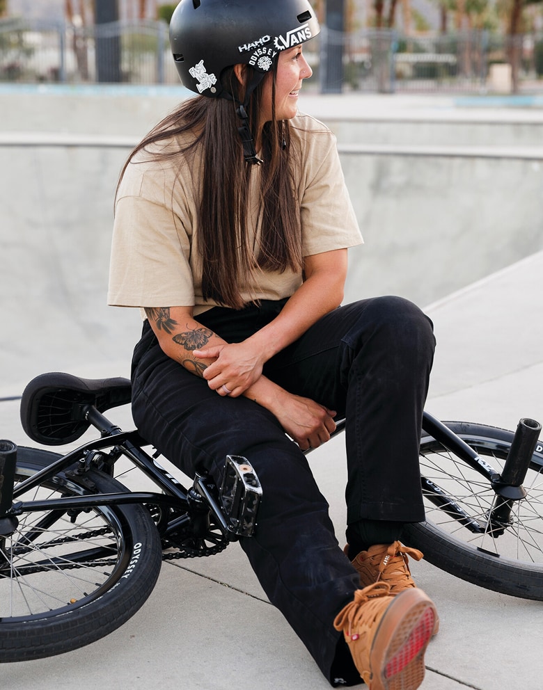 Vans BMX Rider Perris Benegas sits on her bike at a skate park wearing black jeans and the brown BMX Proof WaffleCup.