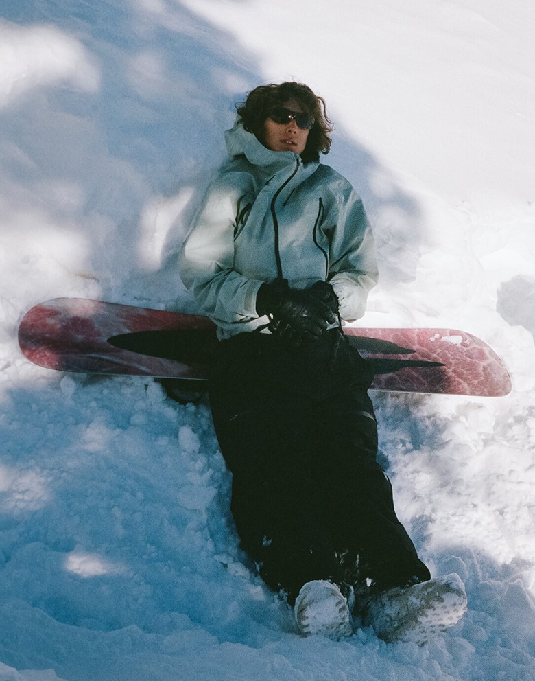 A snowboarder lies in the powder holding his snowboard and wearing Vans snowboarding clothes and boots.