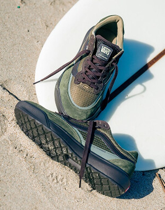 A pair of coal brown UltraRange 2.0 lie on a surfboard at the beach on a sunny day.