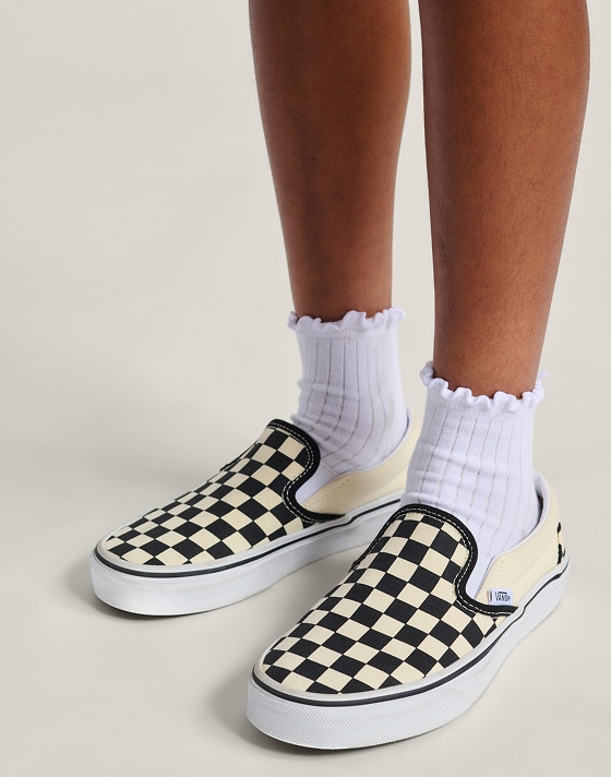 A knee-down shot of a kid wearing black and white Checkerboard Slip-On with white frilly socks in a photography studio.