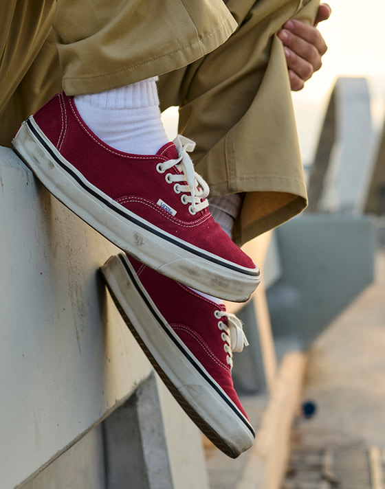 A knee-down shot of a man wearing burgundy Authentic with brown chinos on a bridge during golden hour.