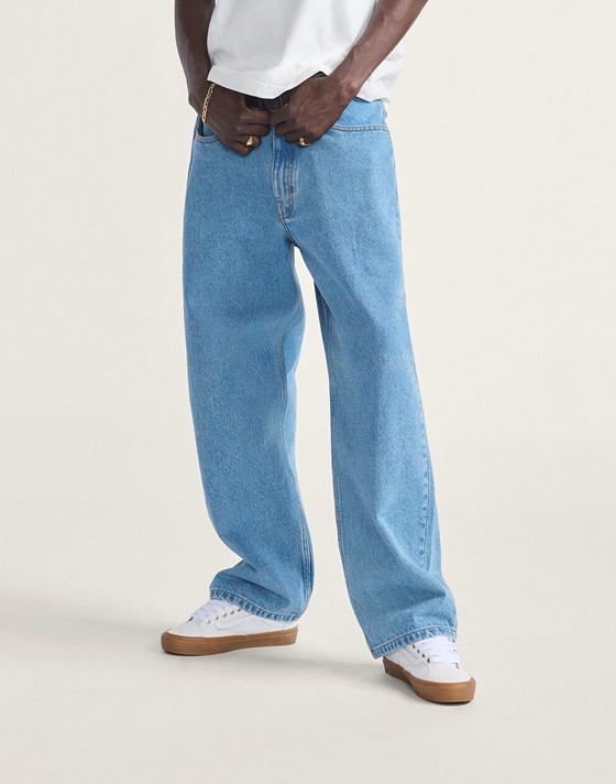 A young man wears blue Check-5 Baggy Denim Pants with white Old Skool and a white t-shirt in a photography studio.