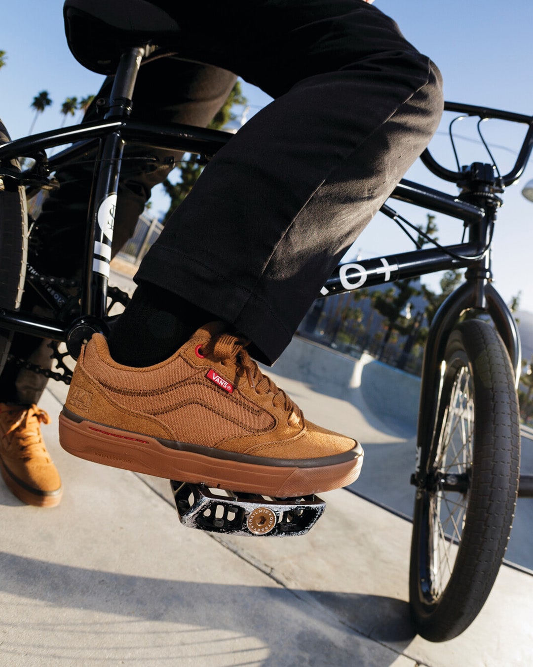 BMX rider at a skate park, standing on this bike, foot resting on the pedals of their bike.