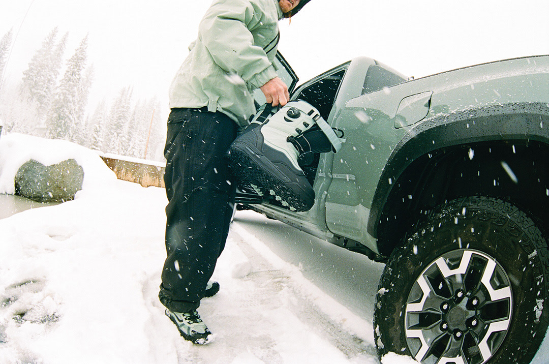 A persons standing outside their truck putting on their snowboard boots.