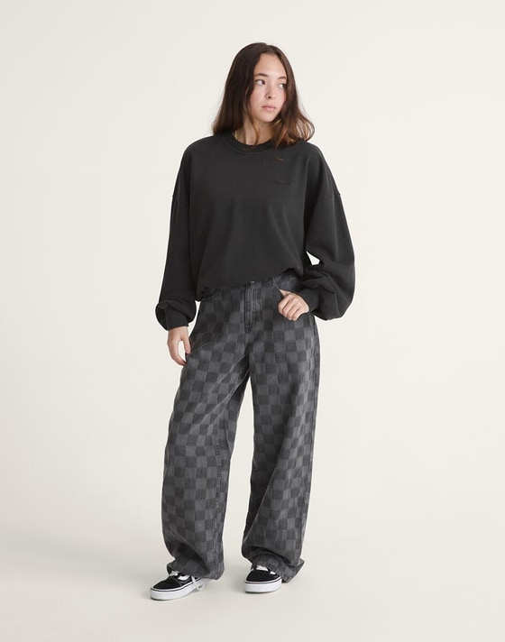 A young woman poses in a photography studio wearing black and gray Checkerboard pants, a black Script Logo Sweatshirt, and black and white Old Skool.