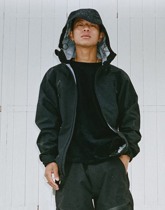 A man stands outside wearing all black with rain on his raincoat looking at the camera.