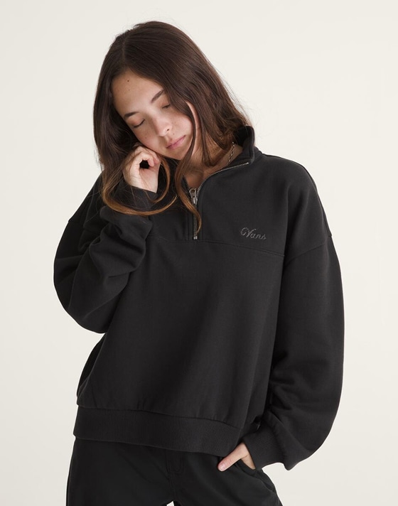 A young woman poses in a photography studio wearing a black Script Logo Sweatshirt, black pants, and pink Old Skool.