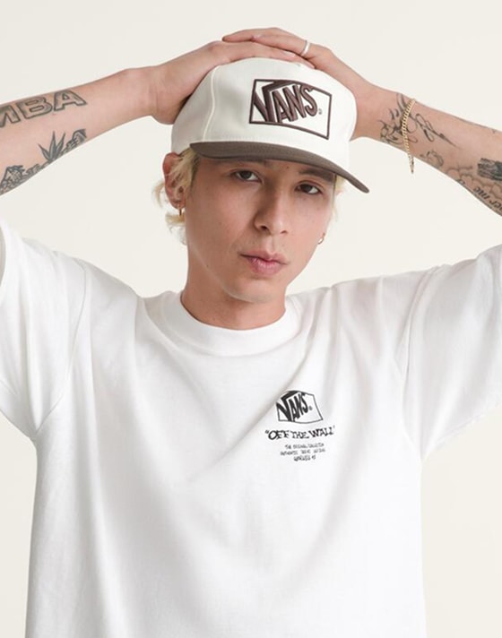 A waist-up shot of a man with bleached blond hair posing in a photography studio wearing a white Off the Wall tee and a white Vans logo hat.
