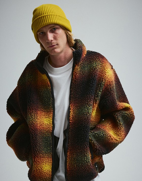 A young man wears the Fleece Sherpa jacket with a yellow beanie and blue jeans in a photography studio.