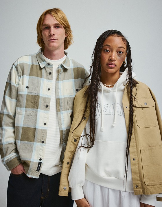 A young man in a Larkspur Plaid Flannel Shirt stands next to a young woman in a tan Drill Chore Coat in a photography studio.
