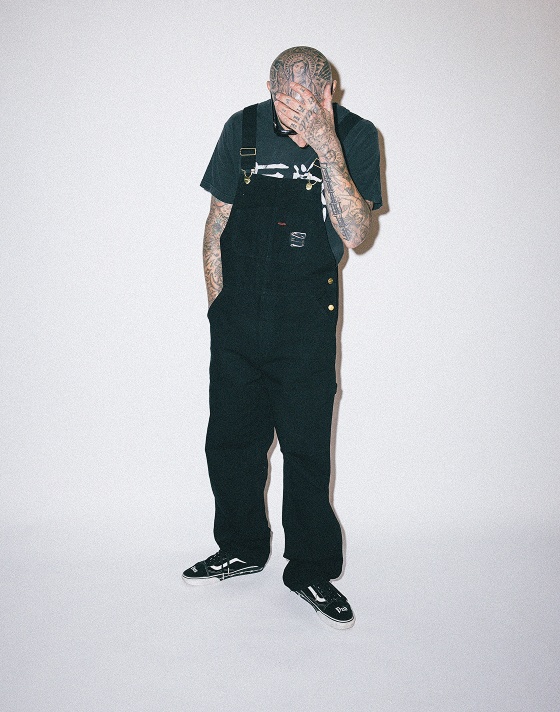 Travis Barker poses wearing black overalls and the Vans x Travis Barker Old Skool in a photography studio.