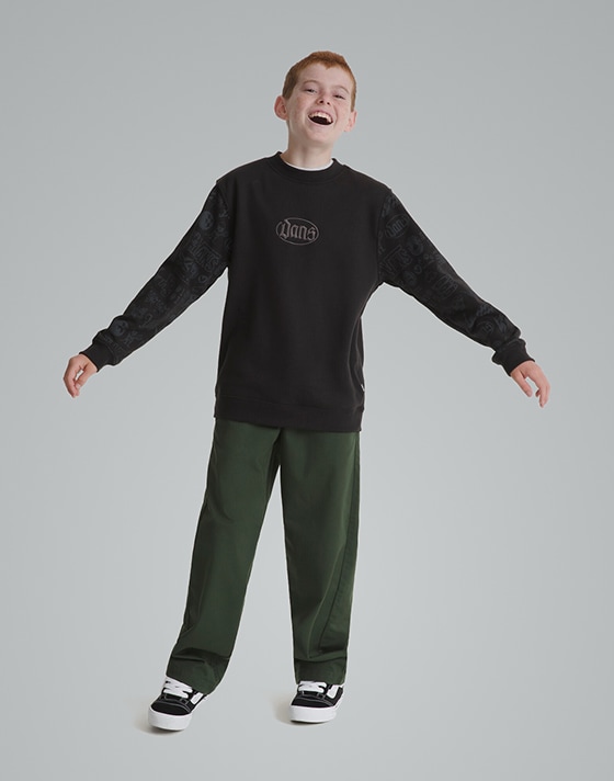 A young boy smiles for the camera wearing a black sweater and black and white Knu Skool in a photography studio.