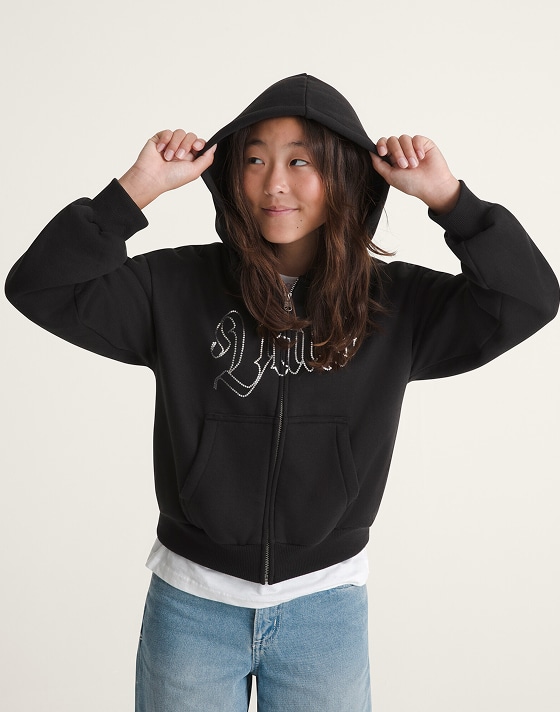 A young girl poses in a Kids Shining Zip Hoodie in a photography studio.s in a photography studio wearing a white Vans “Off the Wall” tee and blue jeans.