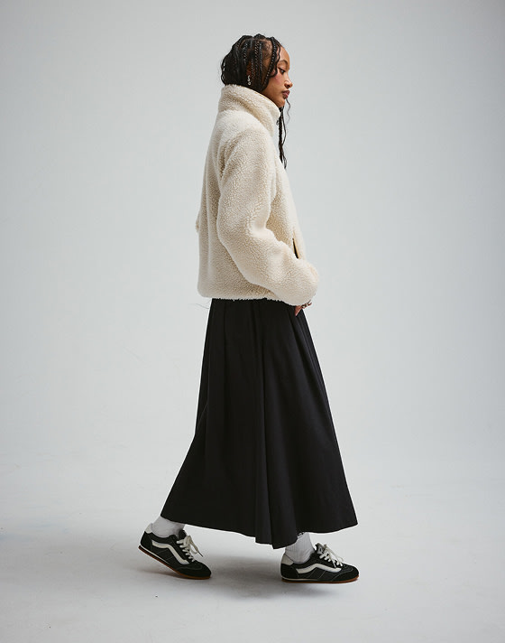 A young woman in a Cozy High Pile jacket and black Super Lowpro walking in a photography studio.