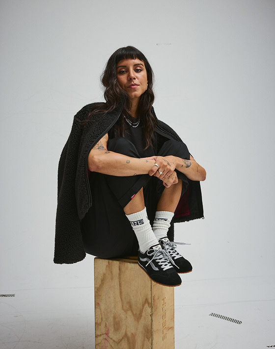A young woman sits on a wooden box wearing Vans clothes and black and Super Lowpro.