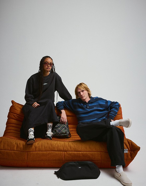 A young man and woman sit on an orange couch wearing beige Slip-On and black Super Lowpro in a photography studio.